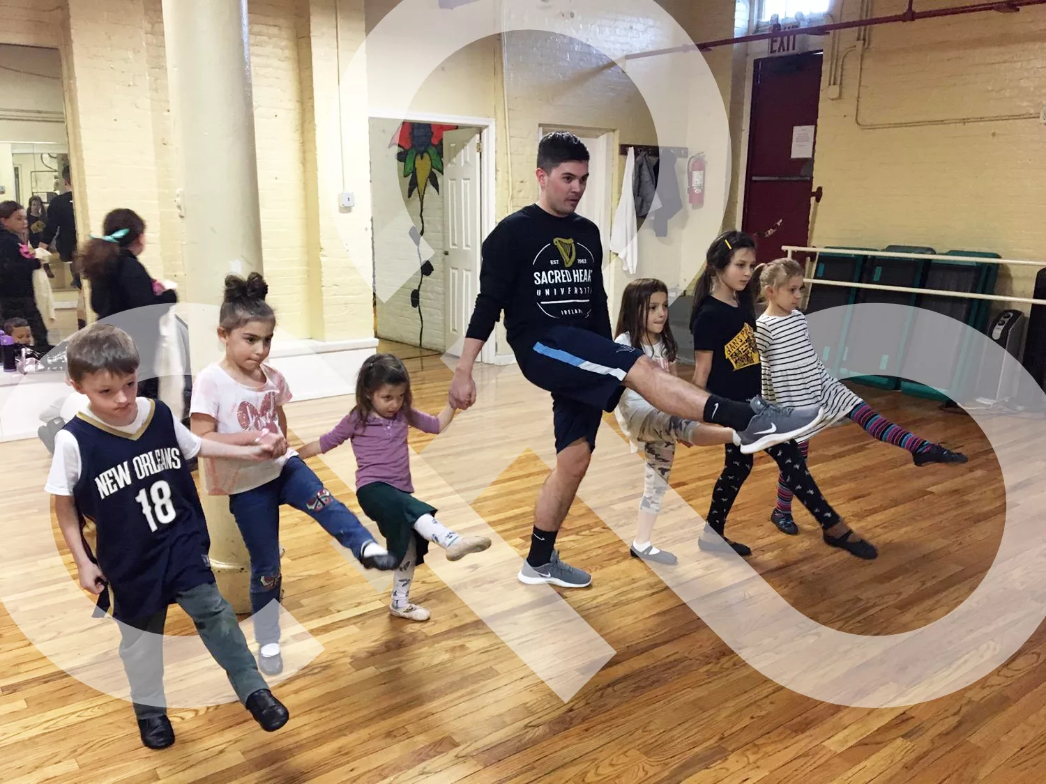 A group of young kids learning basic steps with their dance teacher in a studio with a wooden floor. The IDTANA logo is woven throughout.
