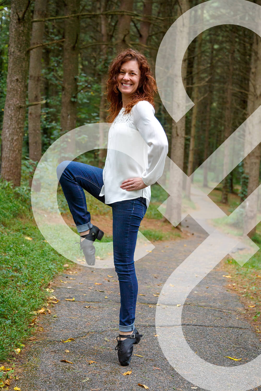 An image of an adult woman dancing in the woods. The woman has red curly hair and is wearing a white shirt, blue jeans, and Irish dance hard shoes. She is balancing on her back toe with her front leg lifted. The IDTANA logo is entwined throughout.